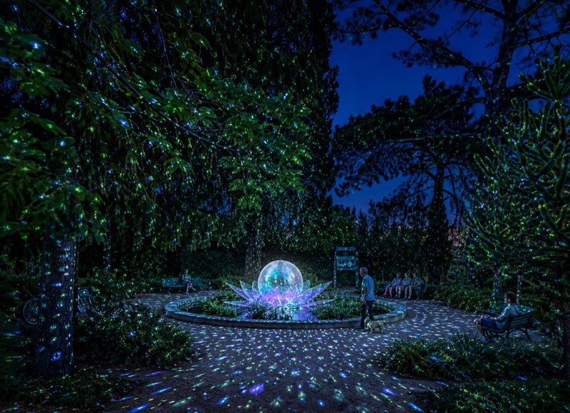 Une fleur de lumière au centre d’un bassin, sous des arbres constellés de points lumineux